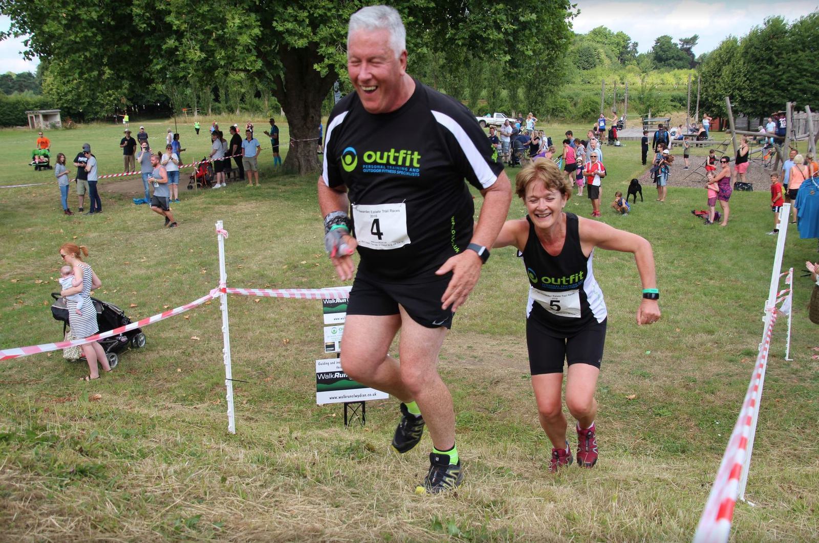Two runners, wearing number 4 and 5 bibs and black athletic gear, are smiling as they ascend a grassy hill during a race. The participant in the front is a man, closely followed by a woman. Spectators and other runners are visible in the background on the sunny day.