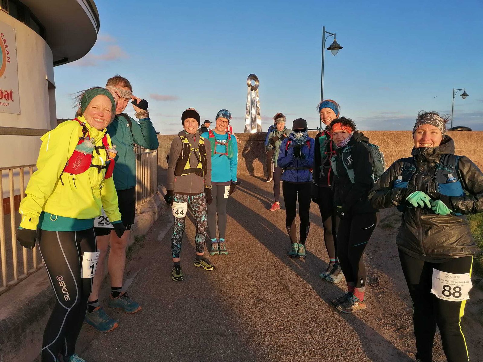 A group of nine people dressed in athletic gear are standing on a paved pathway, smiling and posing for the photo. They have race bibs attached to their clothes. In the background, there's a coastline with a building on the left and a distinctive sculptural landmark.