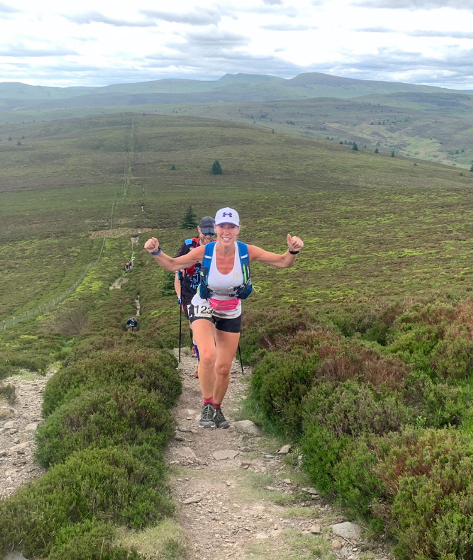 A runner with a white cap and tank top, wearing a race bib number 112, confidently climbs a scenic but rugged trail through green hills under a partly cloudy sky. She raises both arms in a victorious gesture while other participants follow behind.
