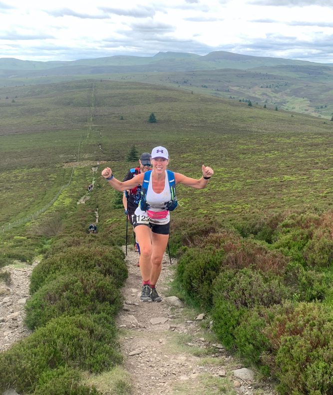 A runner with a white cap and tank top, wearing a race bib number 112, confidently climbs a scenic but rugged trail through green hills under a partly cloudy sky. She raises both arms in a victorious gesture while other participants follow behind.