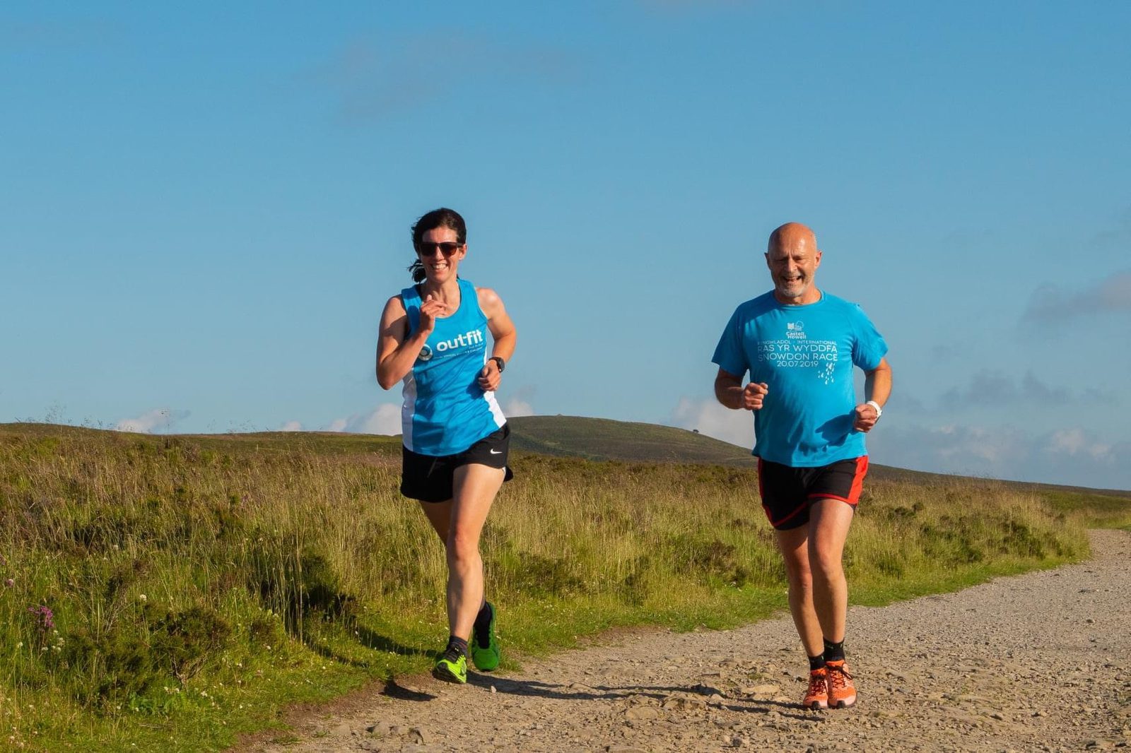 Two people are running on a gravel path through a hilly, rural landscape. The person on the left is wearing a blue tank top and black shorts, and the person on the right is in a blue T-shirt and black shorts with red trim. Both appear to be enjoying the sunny weather.
