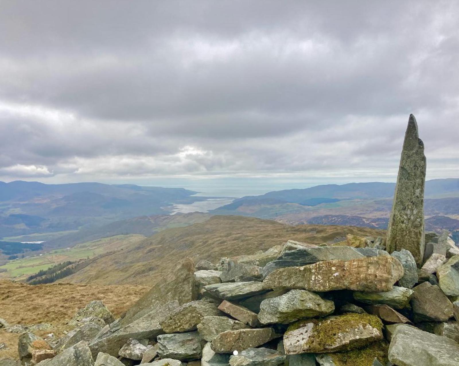 A stone cairn with a tall vertical slab stands on a rocky hilltop under a cloudy sky. The view overlooks a vast landscape of rolling hills and a distant body of water.