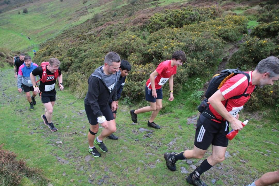 A group of runners, wearing race numbers, ascend a grassy and rocky hill path during an outdoor race. Some wear backpacks and athletic gear, while the surrounding landscape features green bushes and sloping terrain. The sky appears overcast.