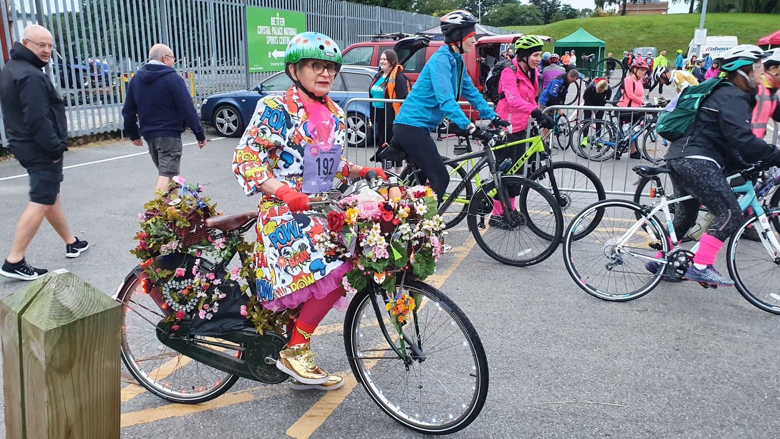A person in a colorful outfit and green helmet stands beside a bicycle adorned with flowers and foliage. Other cyclists, also dressed in bright clothes and helmets, gather around the fenced area. Pedestrians are seen walking by in the background near an event setup.