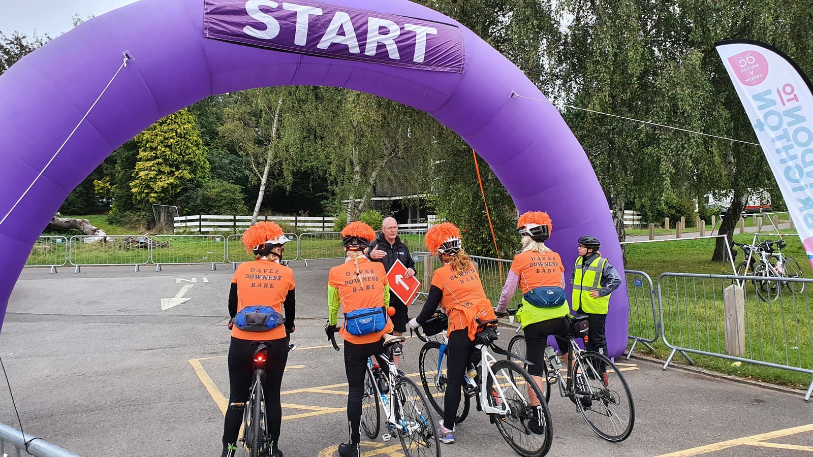 A group of cyclists in bright orange vests gather under a purple inflatable arch that reads "START." One person in a yellow vest stands nearby. The background includes trees, a grassy area, and a fenced enclosure. A flag promoting an event is on the right.