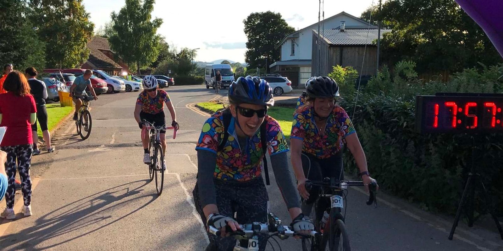 Three cyclists wearing colorful jerseys and helmets cross the finish line, with a digital clock displaying 17:57 in the background. People on the side cheer them on. The scene is set in a rural area with trees, parked cars, and buildings visible.