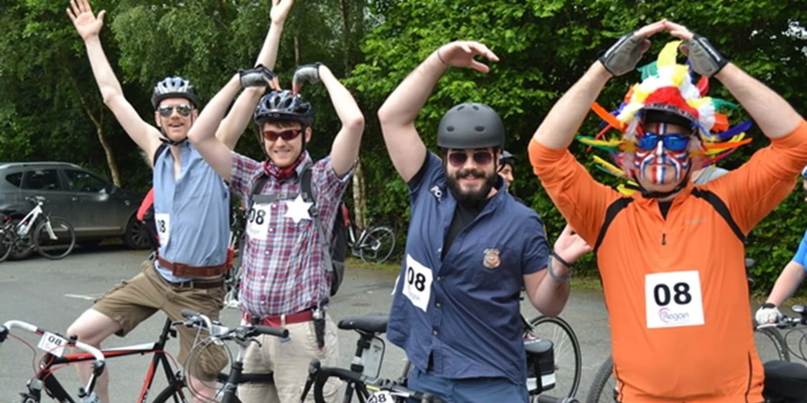 Four people stand with bicycles, all wearing helmets and numbered tags. They energetically pose with their hands in the air. One person is notably dressed in an orange outfit with a colorful feathered headdress and face paint. Trees are visible in the background.
