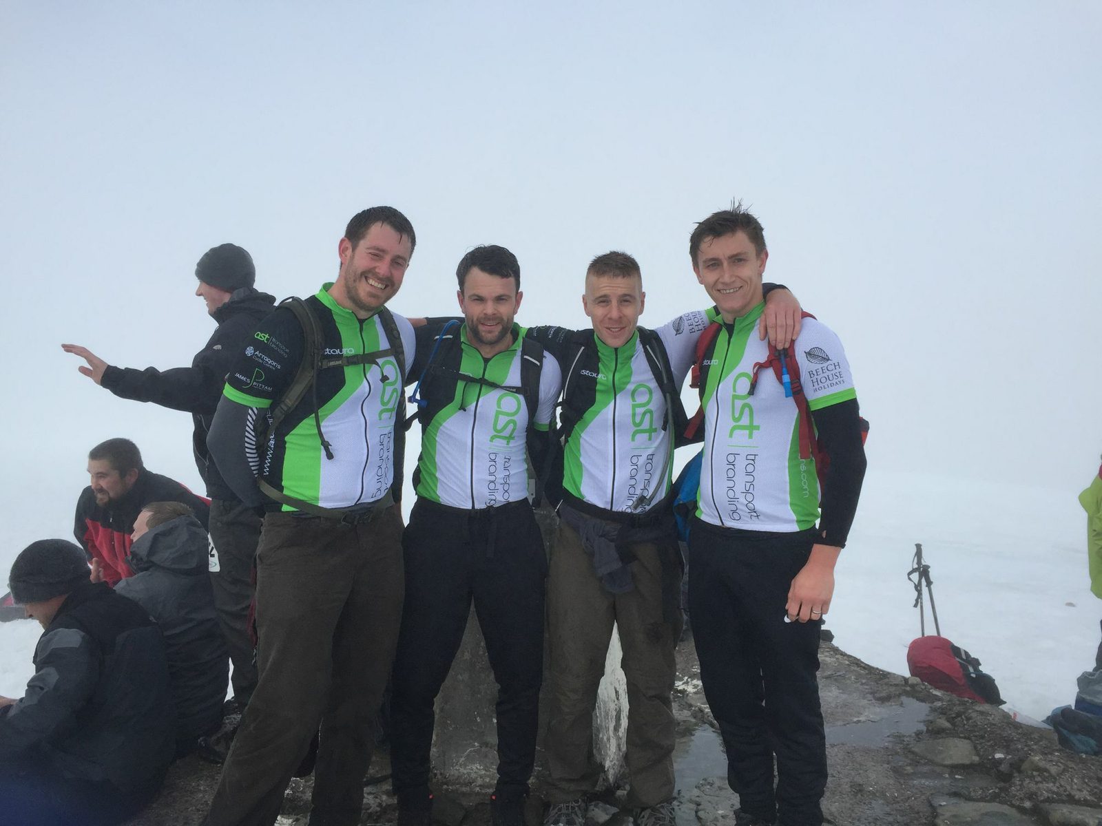 Four men in matching green and white cycling jerseys pose together on a foggy mountain summit. They are smiling and appear happy to have reached the peak. Other climbers in dark clothing are sitting and standing in the background.
