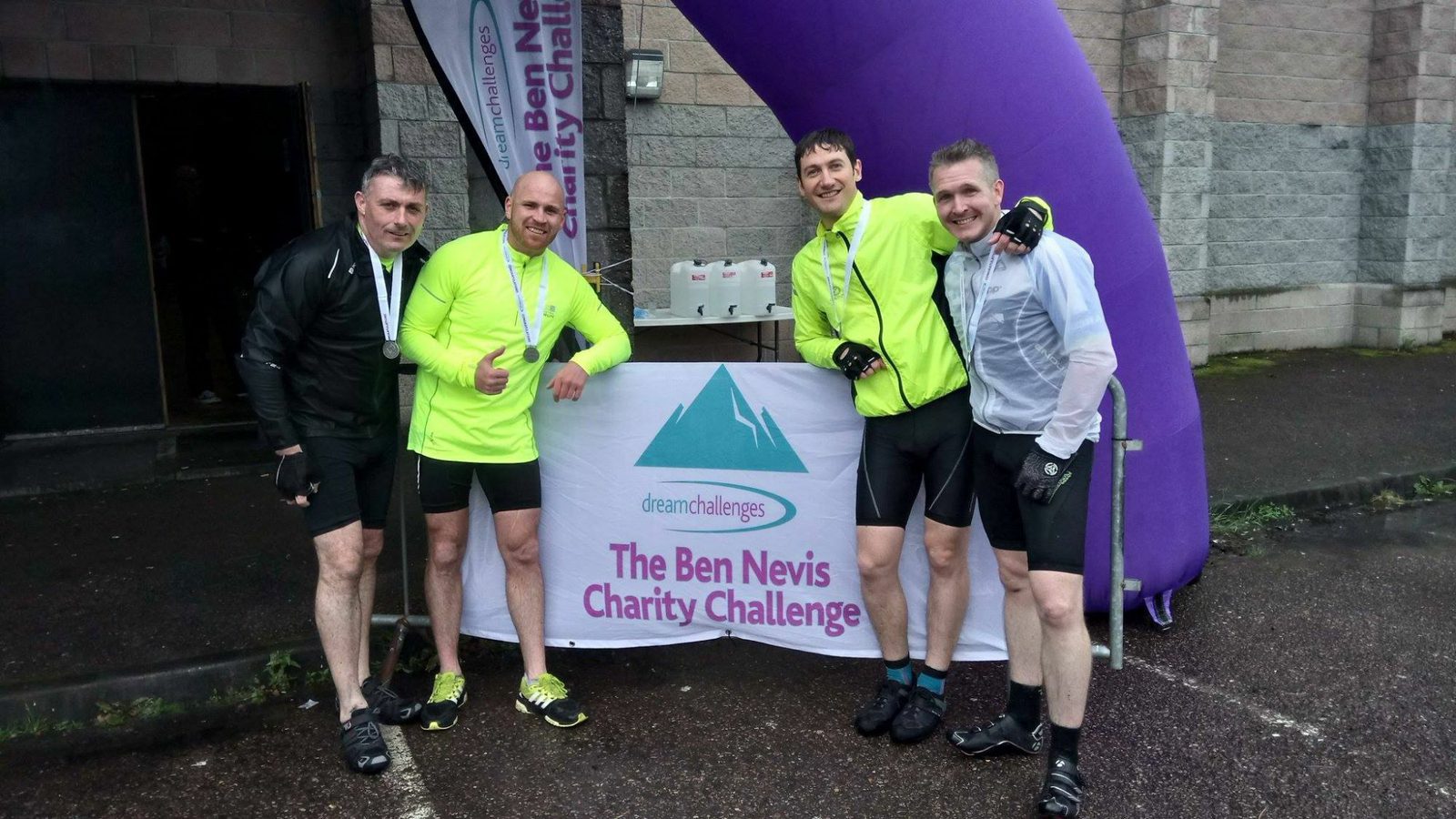 Four men in cycling gear pose with medals around their necks at the finish line of "The Ben Nevis Charity Challenge." They stand under an arch and behind a banner displaying the event's name and logo, showing accomplishment and camaraderie. The ground is wet, indicating recent rain.