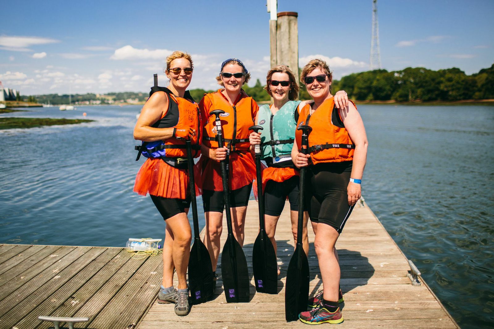 Four women stand close together on a dock, all dressed in orange life jackets and athletic gear, each holding a paddle. The background features a calm body of water, greenery, and a partly cloudy sky. The women are all smiling, appearing happy and relaxed.