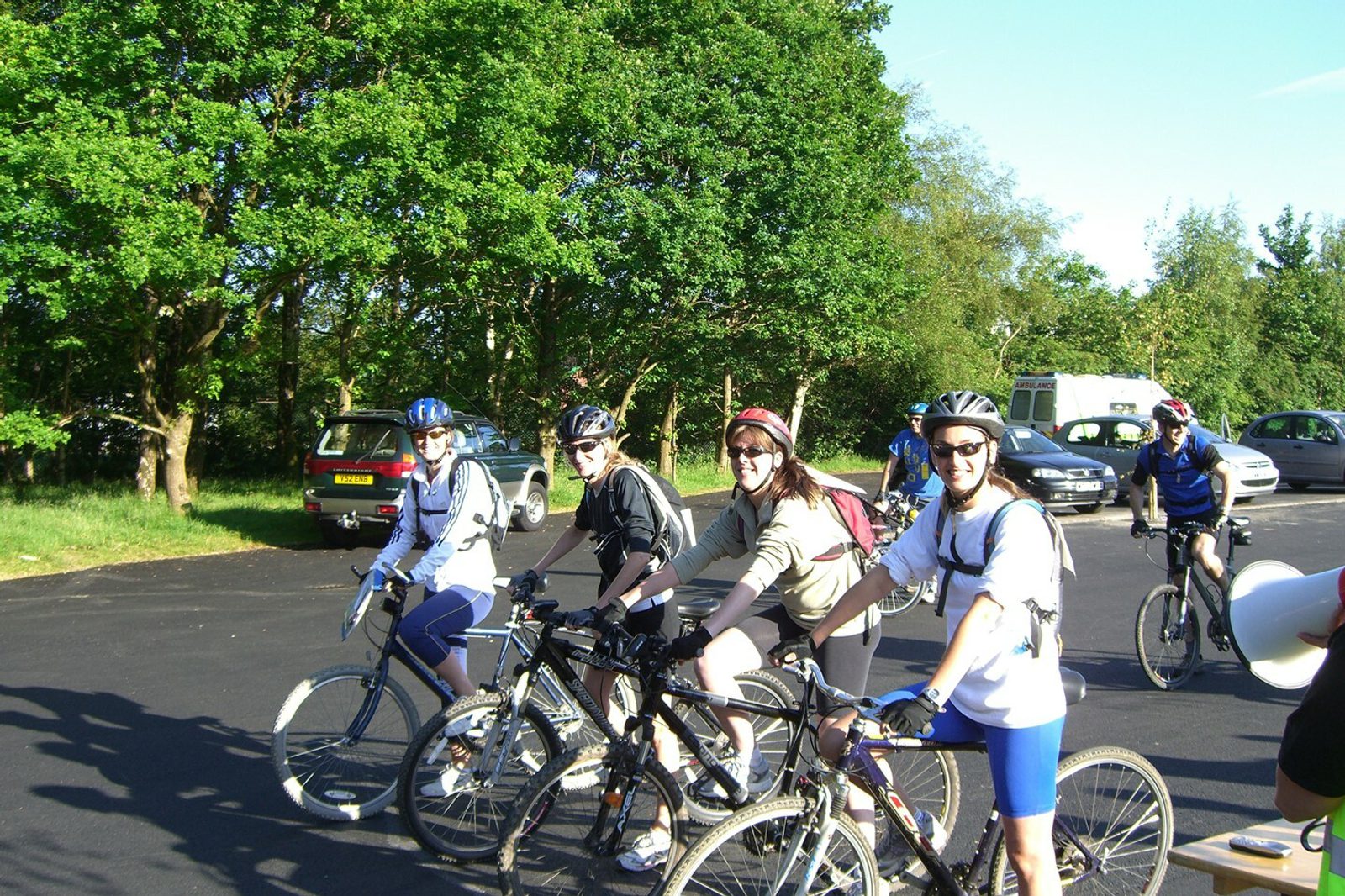 A group of four cyclists posing with their bicycles in a parking area surrounded by trees. They are wearing helmets, sunglasses, and casual cycling attire. Several parked cars and additional cyclists can be seen in the background, along with a bright, clear sky.