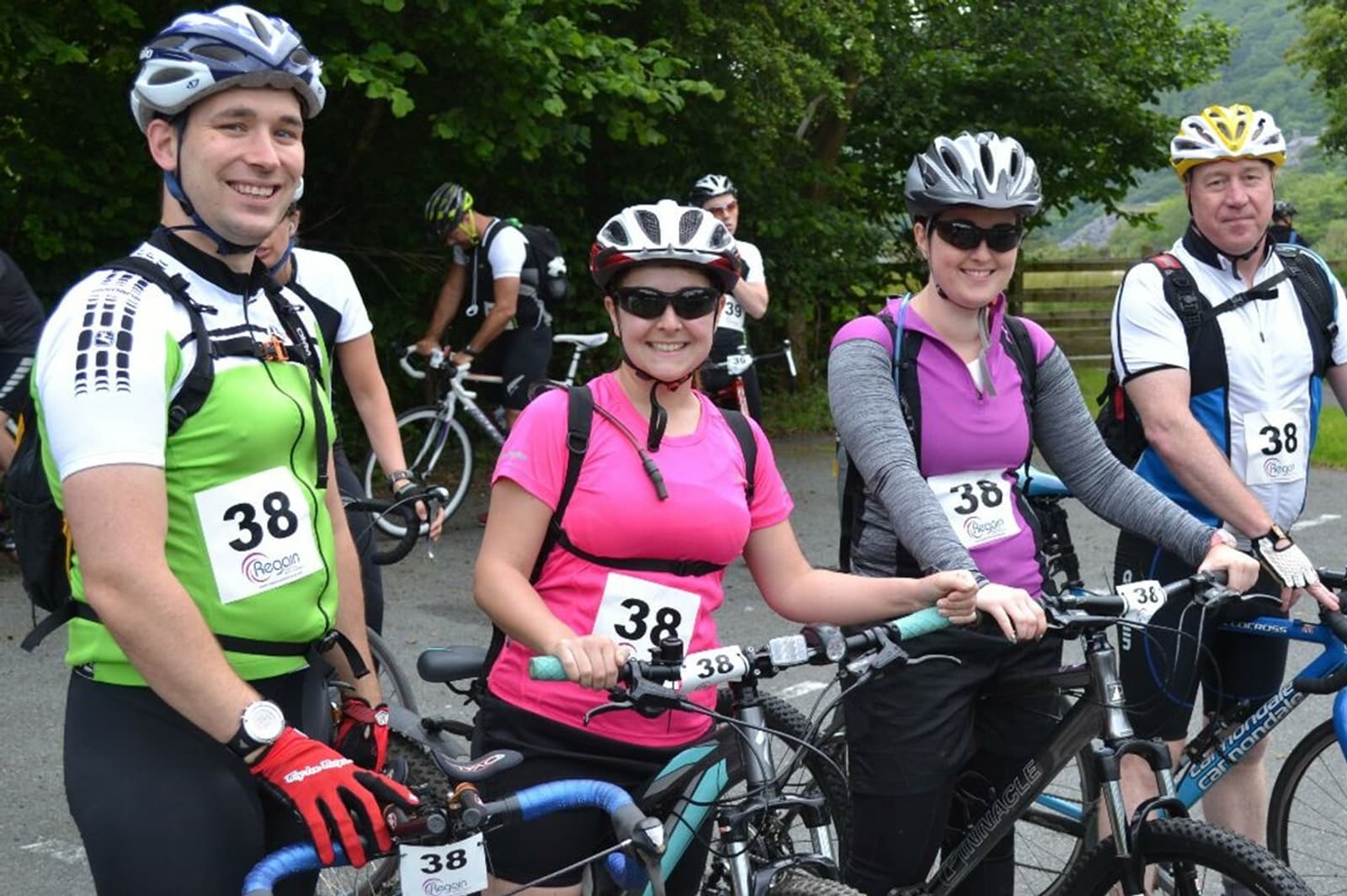 Four cyclists wearing helmets and numbered bibs, standing with their bikes on a road surrounded by green foliage. The three in the foreground are smiling and have their hands on their handlebars, while the fourth person is slightly behind, looking at the camera.