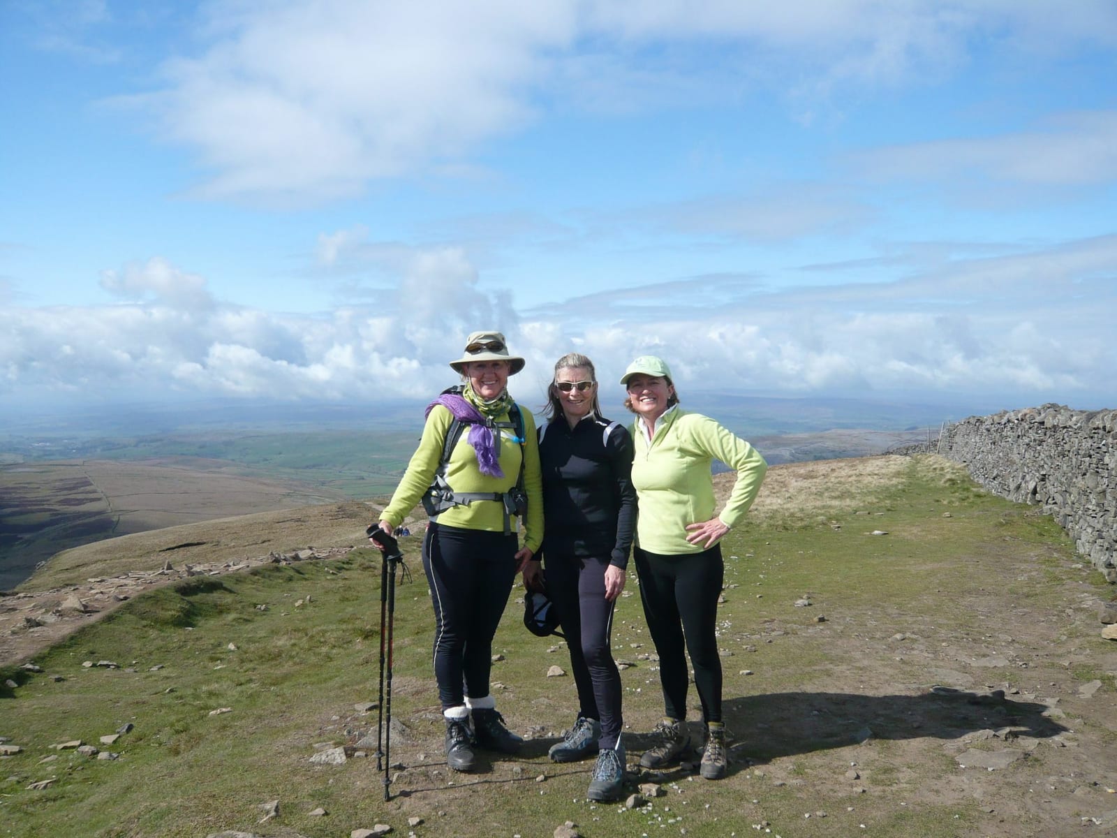 Three women dressed in hiking gear stand together on a mountain trail. They are smiling at the camera, with a scenic backdrop of rolling hills, a stone wall, and a partly cloudy sky. The terrain is grassy and appears to be at a high elevation.