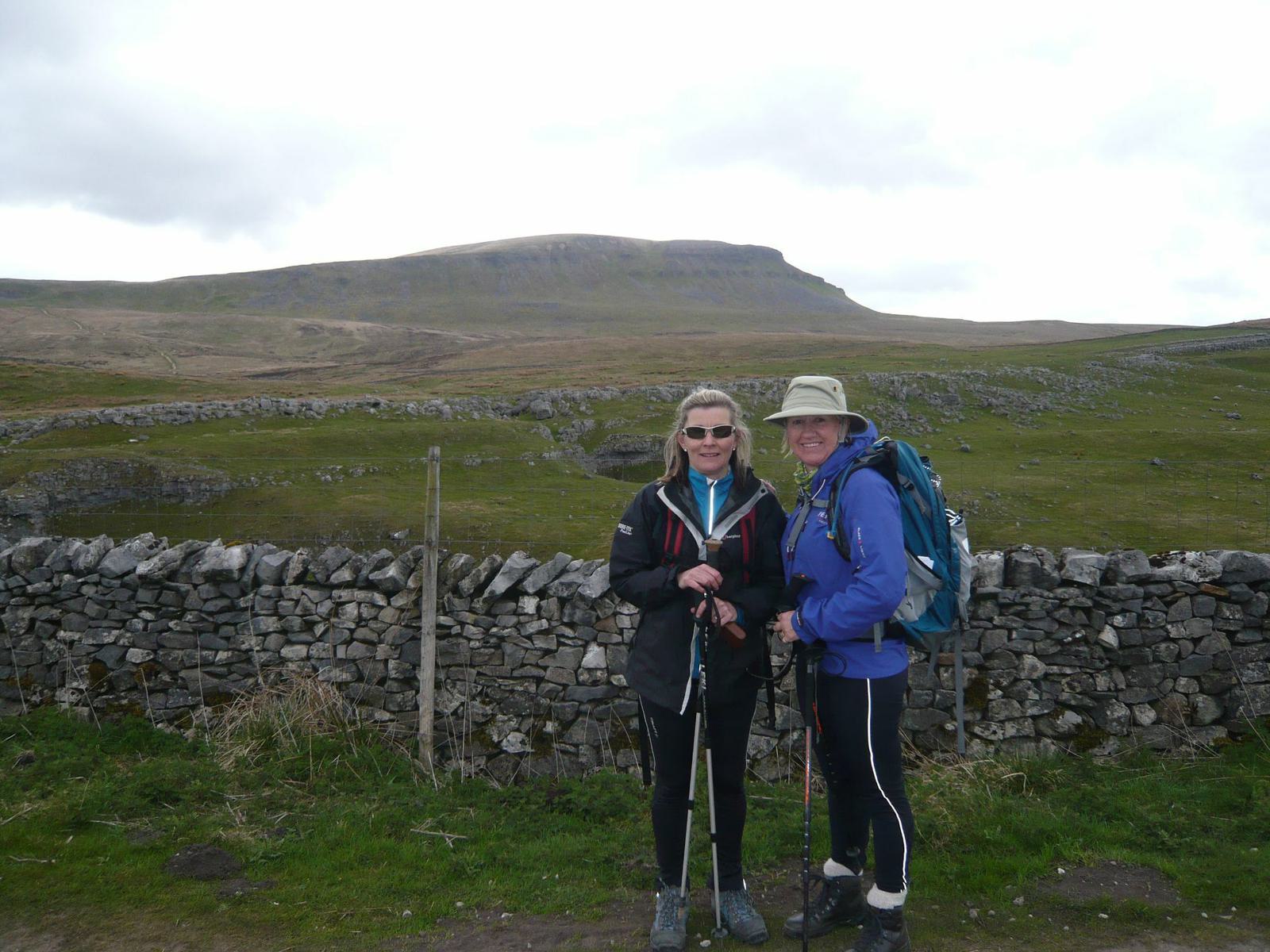 Two hikers stand side by side, smiling and holding trekking poles, in front of a stone wall. One wears a blue jacket and hat with a backpack; the other wears a black jacket. Behind them, rolling green hills, rocks, and a large hill under a cloudy sky are visible.