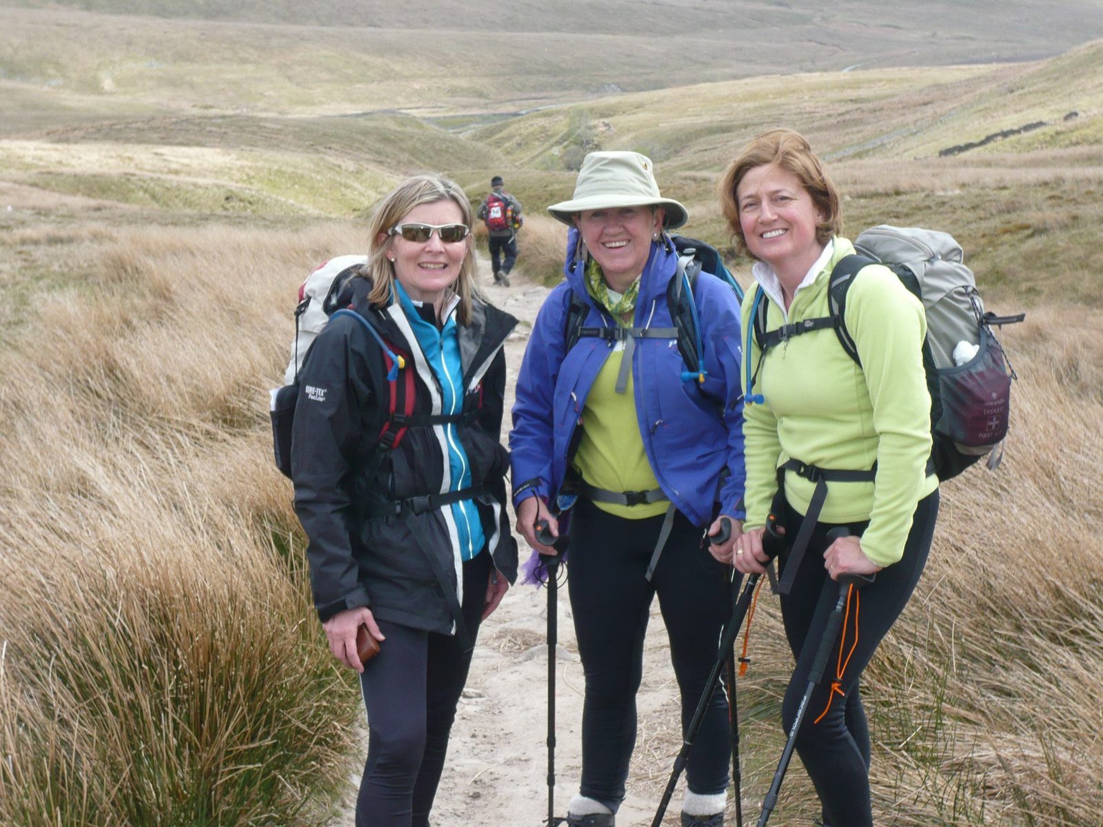 Three women stand on a trail in a grassy, hilly landscape. They are wearing hiking gear, including jackets, backpacks, and using trekking poles. They are smiling and appear to be enjoying their outdoor adventure. A person in the background is also hiking.