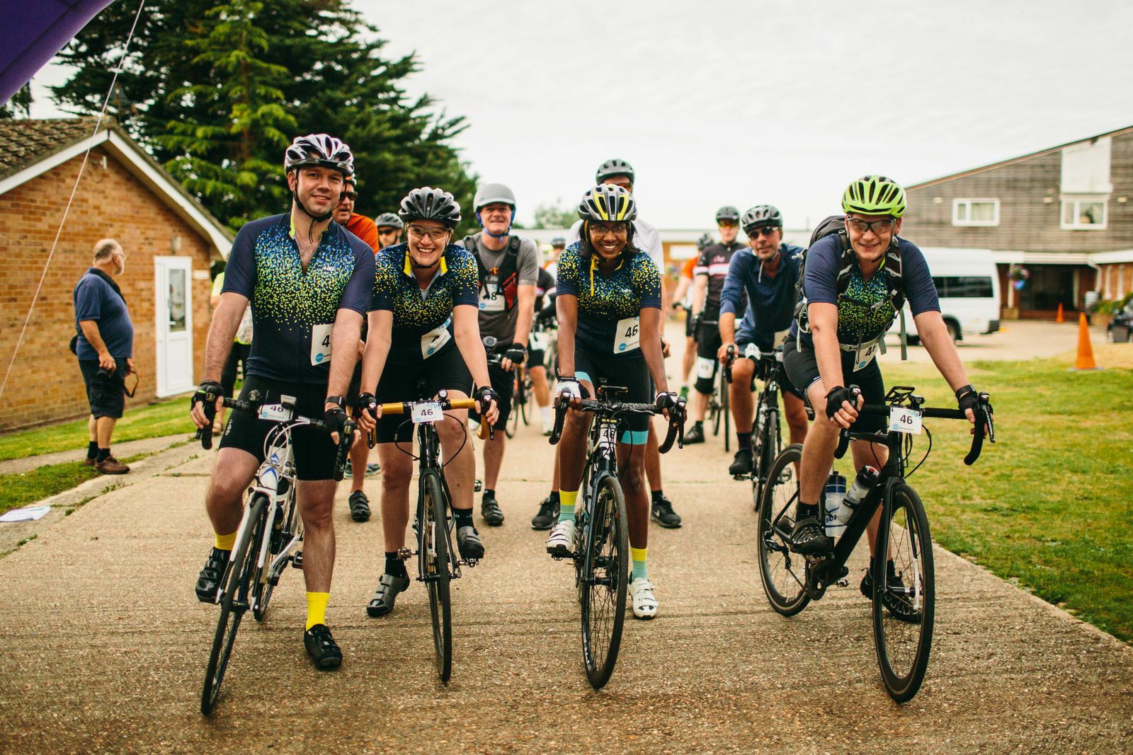 A group of cyclists in matching jerseys and helmets are smiling at the camera as they prepare for a race. They are standing with their bikes on a paved area, with buildings and trees in the background. The atmosphere appears friendly and lively.