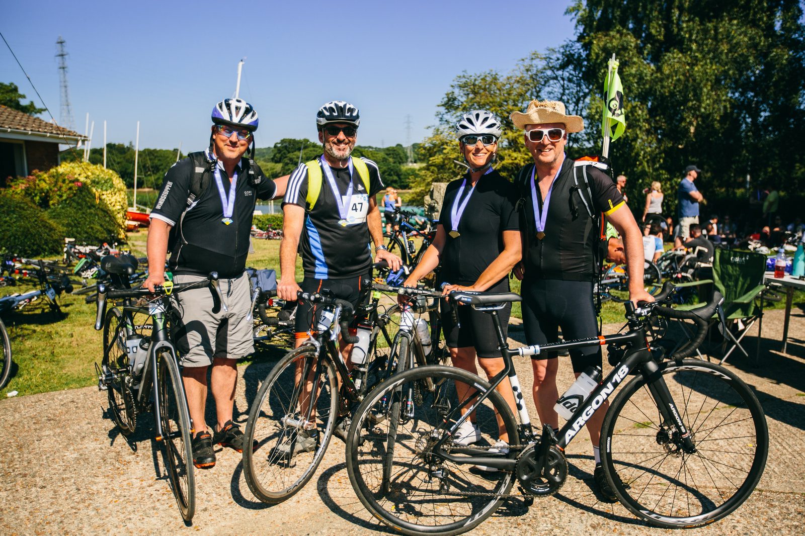 Four cyclists wearing helmets and sports gear stand together smiling, each holding their bikes. They all have medals around their necks, likely celebrating the completion of a cycling event. The background shows other cyclists, bikes, trees, and a green event flag.