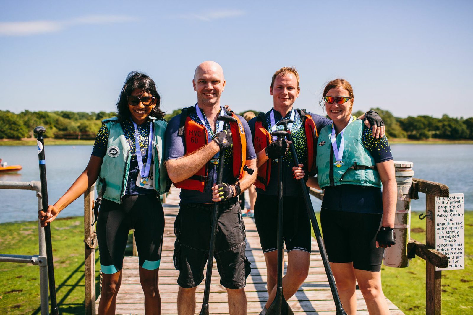 Four people stand on a wooden dock, holding paddles and wearing life vests. They all have medals around their necks and are smiling at the camera. A lake is visible behind them under a clear blue sky.