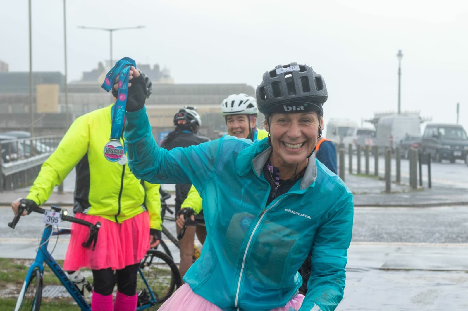A cyclist wearing a blue jacket and helmet happily displays a medal while standing beside their bike. Other cyclists in colorful gear are visible in the background on a wet, overcast day near a city street with parked cars and buildings.