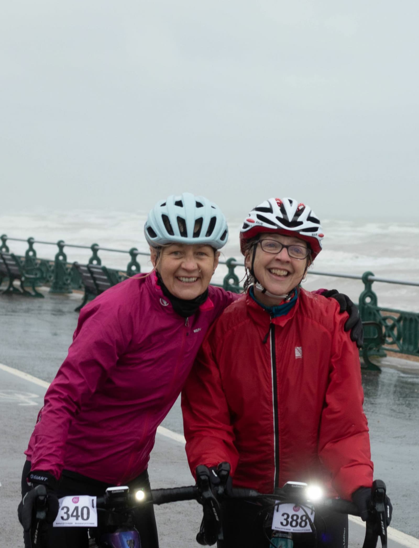 Two smiling individuals wearing helmets and red jackets pose with their bicycles on a rainy beachside promenade. Each has a bike number, 340 and 388. They appear cheerful despite the overcast, wet weather and are standing close to the ocean.