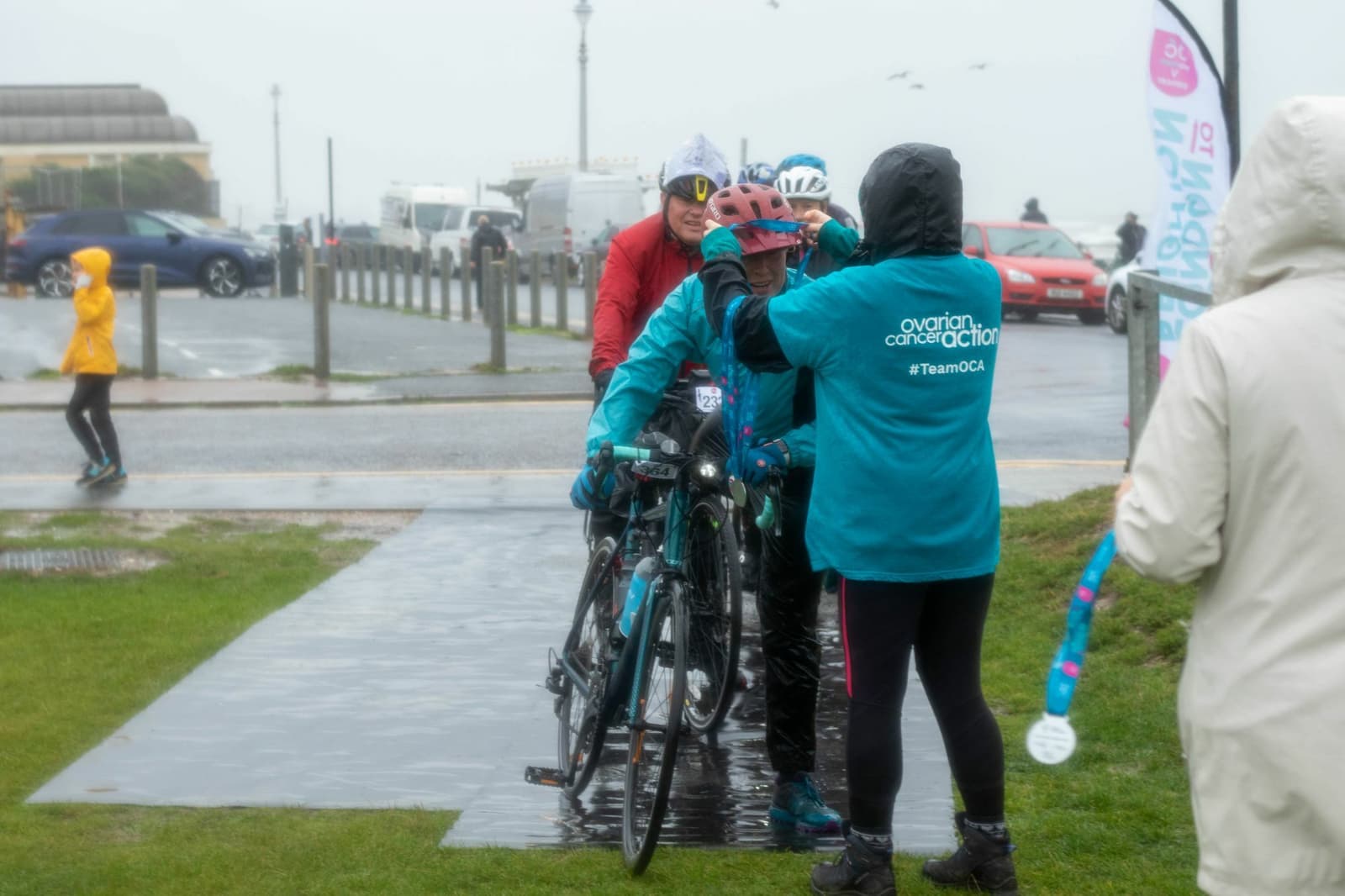 A person wearing a teal "ovarian cancer action" shirt helps another person adjust their helmet in a rainy outdoor setting. Several people in raincoats and bicycles are present. A banner and some cars are visible in the background.