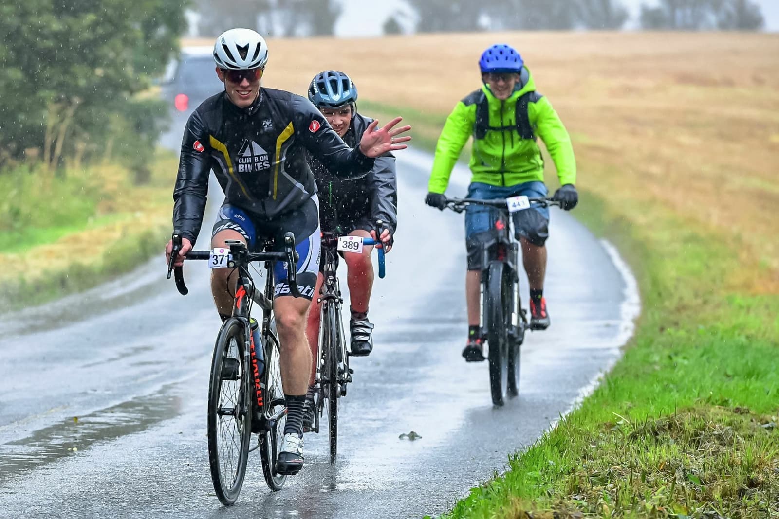Three cyclists ride on a wet, rural road during a rainy day. The foremost cyclist waves at the camera, wearing a black and white outfit and helmet. The other two cyclists follow, wearing rain gear and helmets. The background features grassy fields and trees.