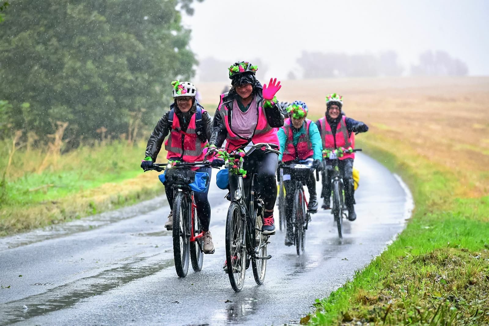 A group of cyclists wearing colorful rain gear and helmets decorated with flowers ride down a wet, rural road on a rainy day. The leading cyclist waves at the camera while the others follow closely behind. Trees and fields line the road.