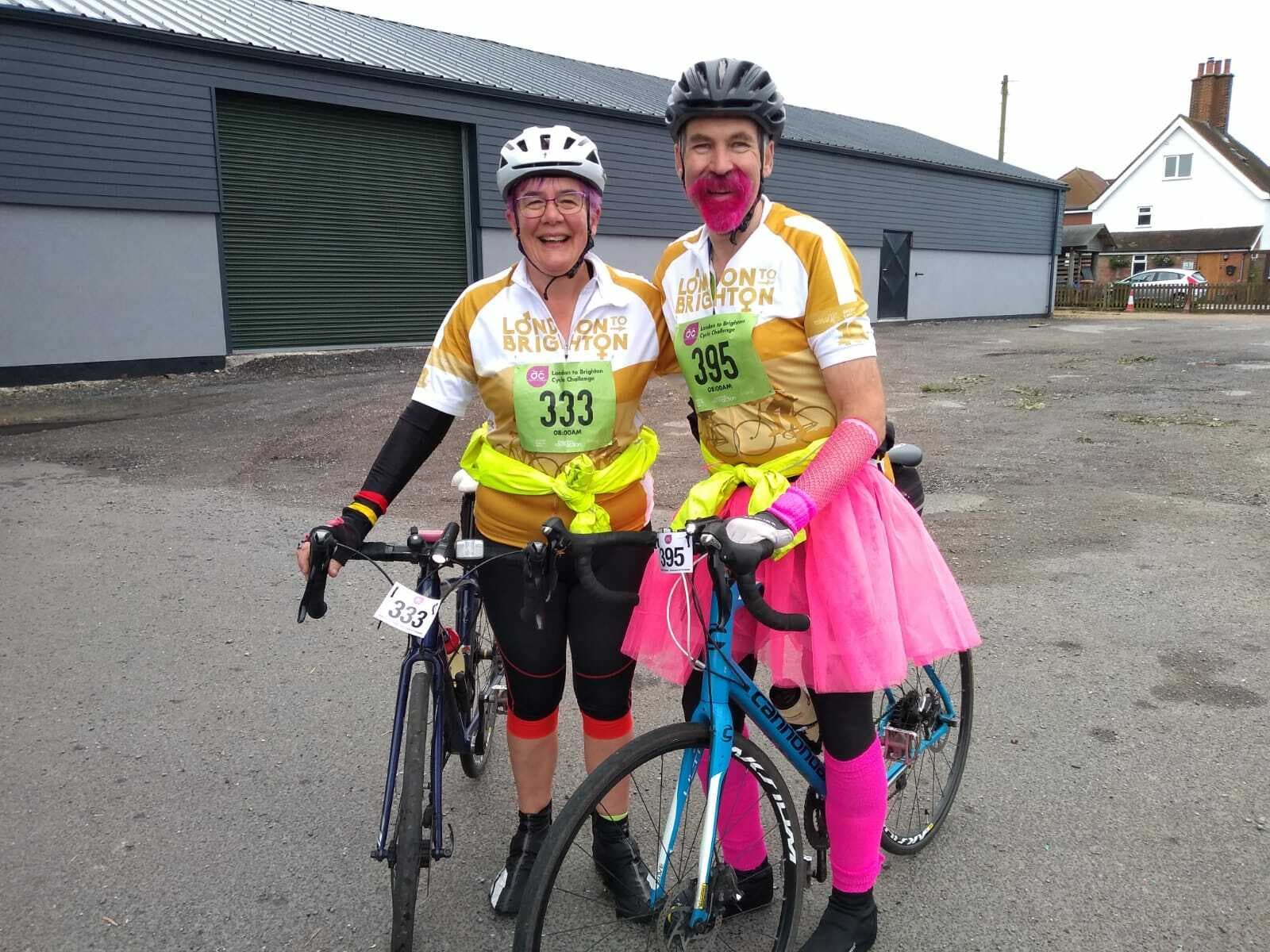Two cyclists pose with their bikes in matching yellow and white "London to Brighton" jerseys and green bibs numbered 333 and 395. One cyclist has a pink beard, pink leg warmers, and a pink tutu, while the other is smiling broadly. They are standing in a paved area.
