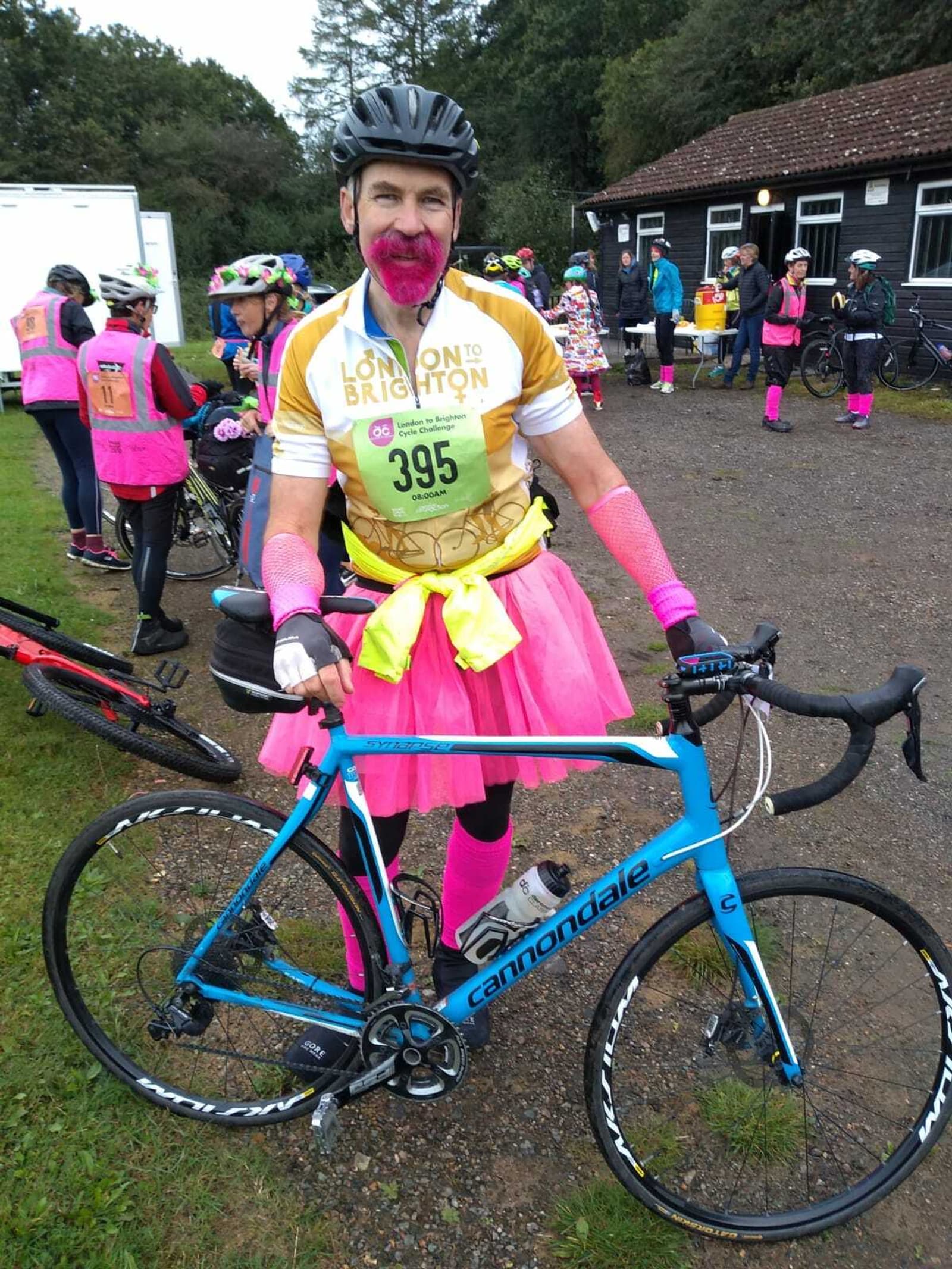 A man in a pink tutu, arm warmers, and socks, wearing a helmet and a "London to Brighton" cycling shirt numbered 395, poses with a blue Cannondale bike. Other cyclists in similar outfits are gathered in the background near a building and some bikes.