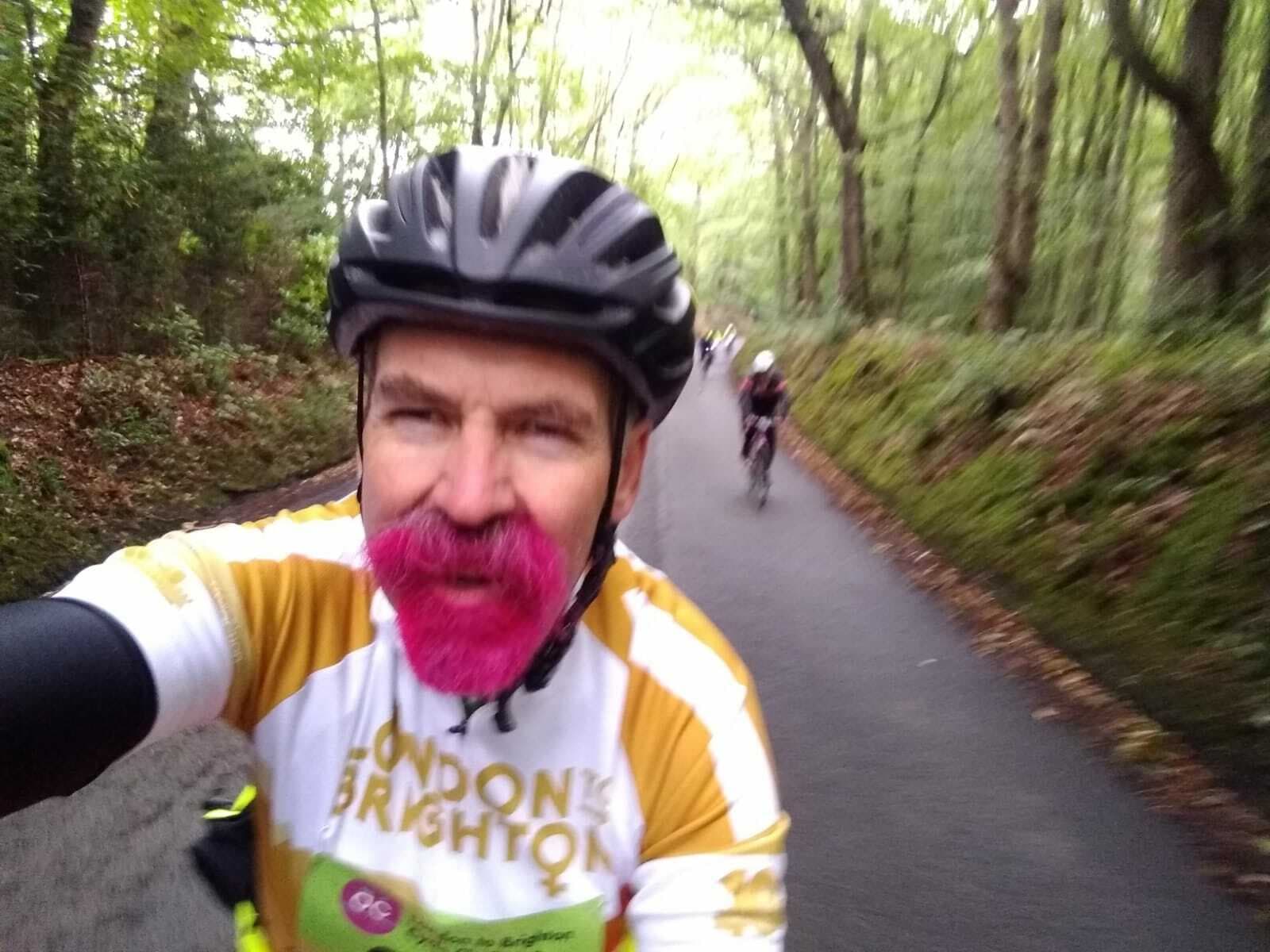 A cyclist with a pink beard and a black helmet is taking a selfie while riding on a tree-lined road. He is wearing a white and yellow "London to Brighton" jersey. Another cyclist is visible in the background. The surrounding area is lush and green.