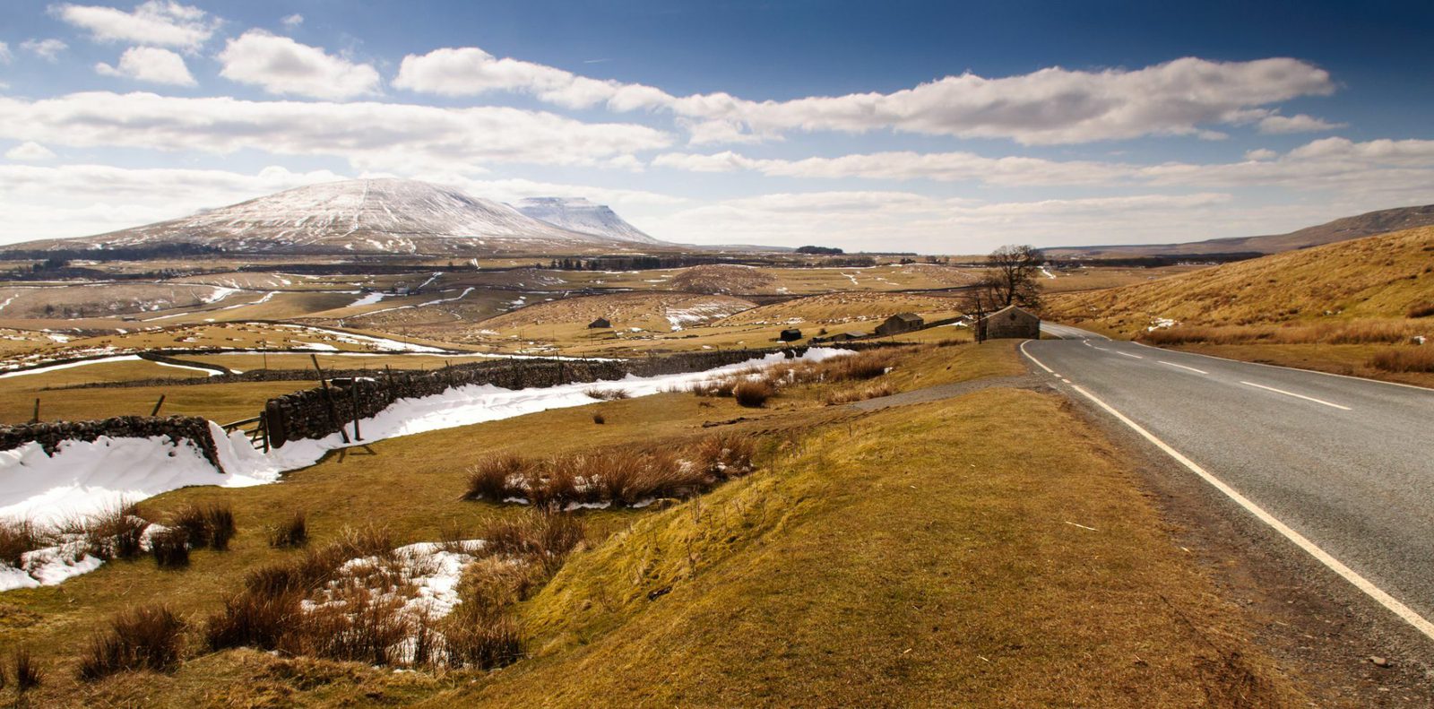 A scenic countryside landscape featuring a winding road, rolling hills, sparse patches of snow, dry stone walls, and a lone tree under a partly cloudy blue sky. Snow-capped hills and mountains are visible in the background.