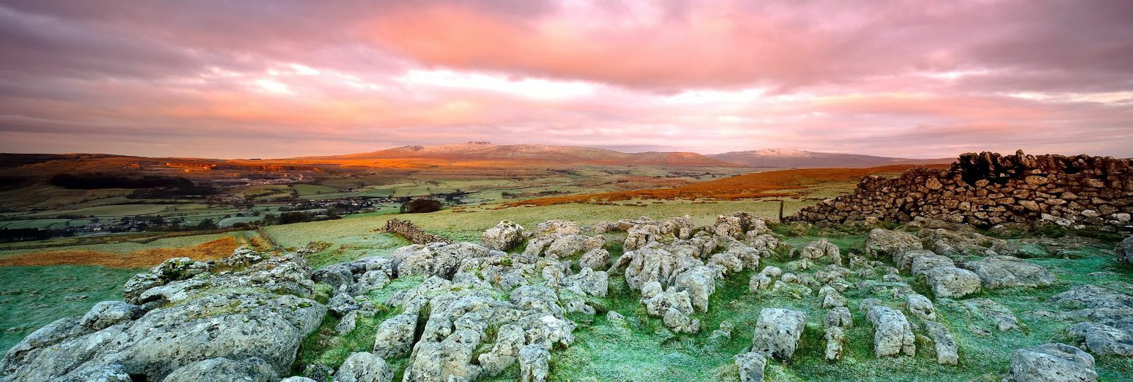 Panoramic view of a countryside landscape at sunrise or sunset, with rocky foreground, rolling hills, a patchwork of fields, and distant mountains under a sky painted with vibrant hues of pink, orange, and purple clouds. Ancient stone walls are scattered throughout.