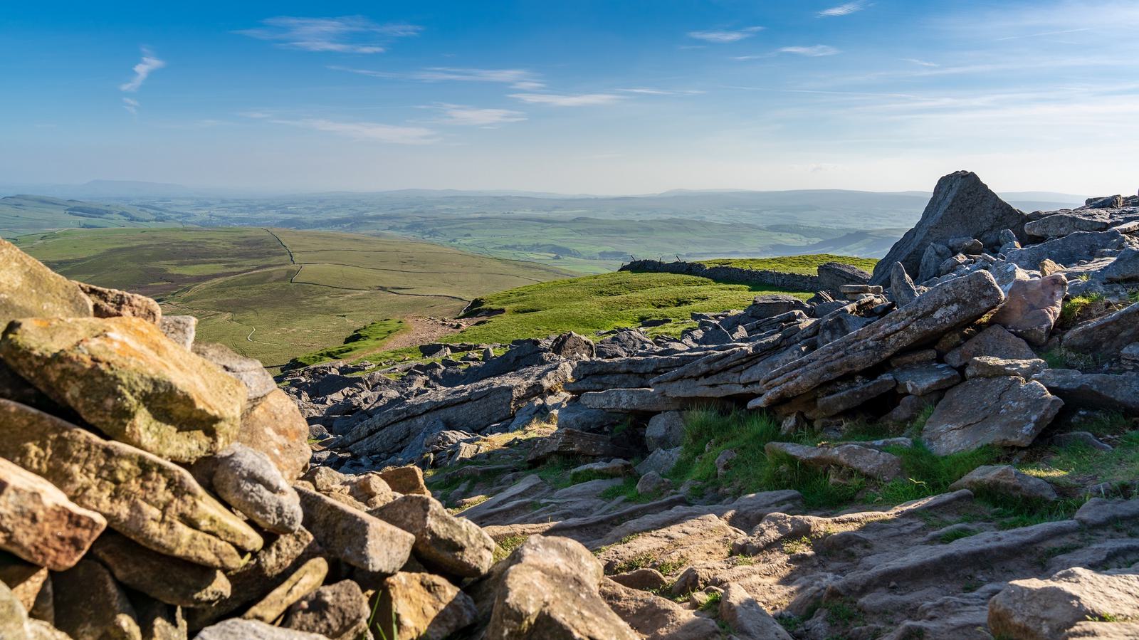 A scenic view from a rocky hilltop overlooking a vast, green countryside with rolling hills under a clear blue sky. The foreground features scattered rocks and boulders, with patches of grass interspersed among them.
