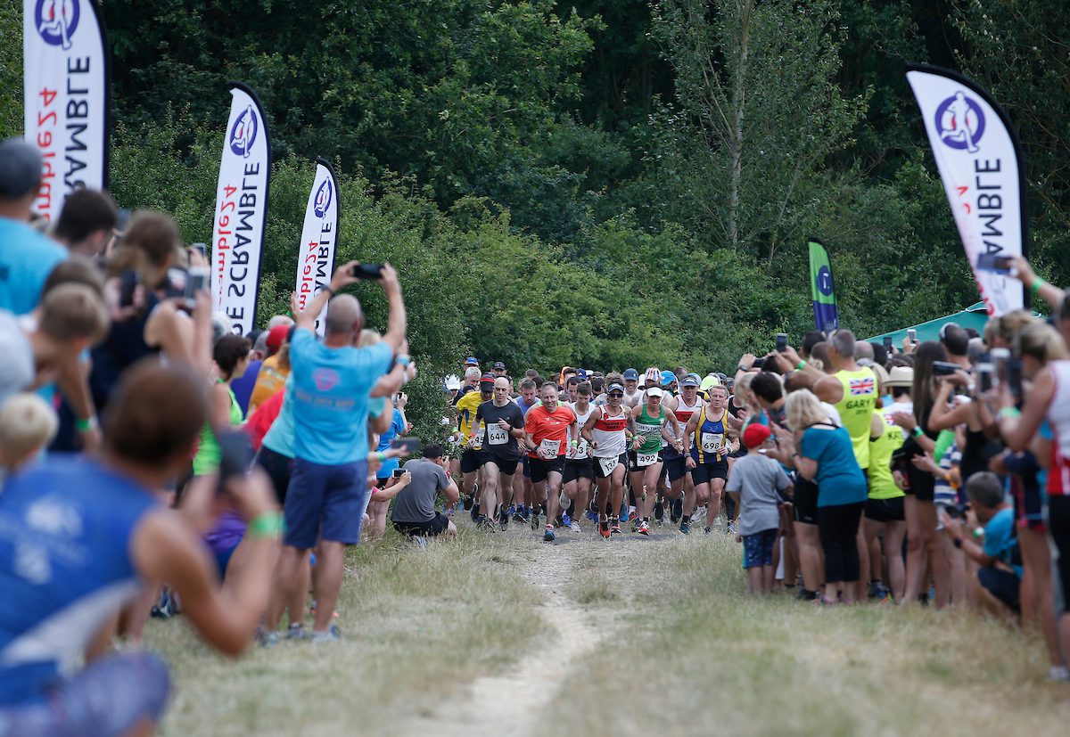 A group of runners starts a race on a grassy path, surrounded by spectators. People line both sides of the path, cheering and taking photos. Trees and various banners are visible in the background.
