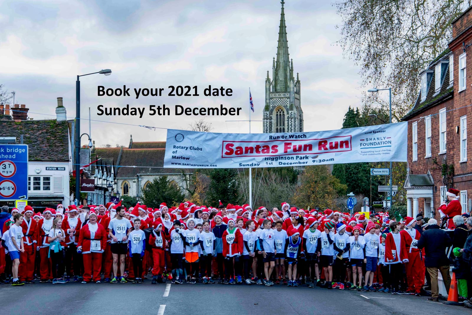 A large group of people dressed in Santa costumes stand at the starting line of a fun run event, with a banner overhead reading "Santas Fun Run" and "Sunday 5th December." Trees, buildings, and a church spire are visible in the background.