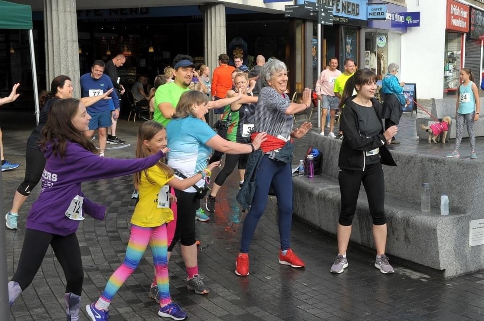 A group of people, including children and adults, are energetically participating in a group dance or warm-up session in an outdoor plaza. They are lined up with smiles, wearing casual sportswear. Some are wearing race bibs, indicating a possible running event.