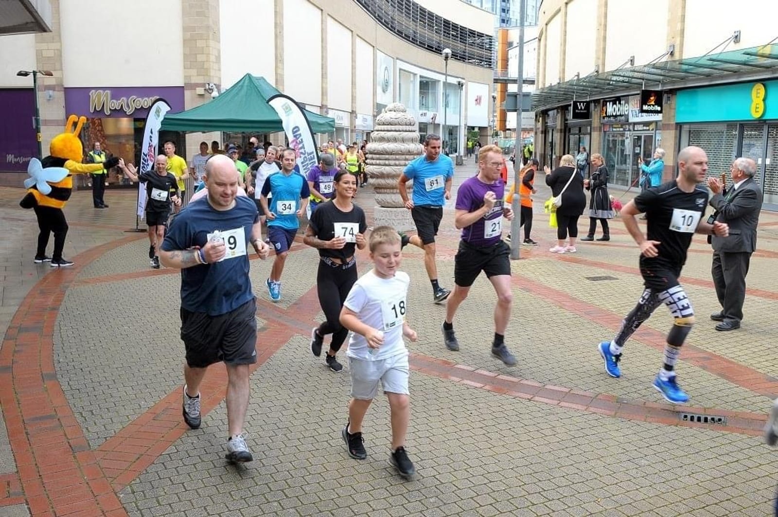 A group of runners participates in a race in an outdoor shopping area. Some are wearing numbered race bibs, and there is a variety of athletic clothing. Spectators and volunteers can be seen along the sides, and there is a person in a large yellow mascot costume in the background.