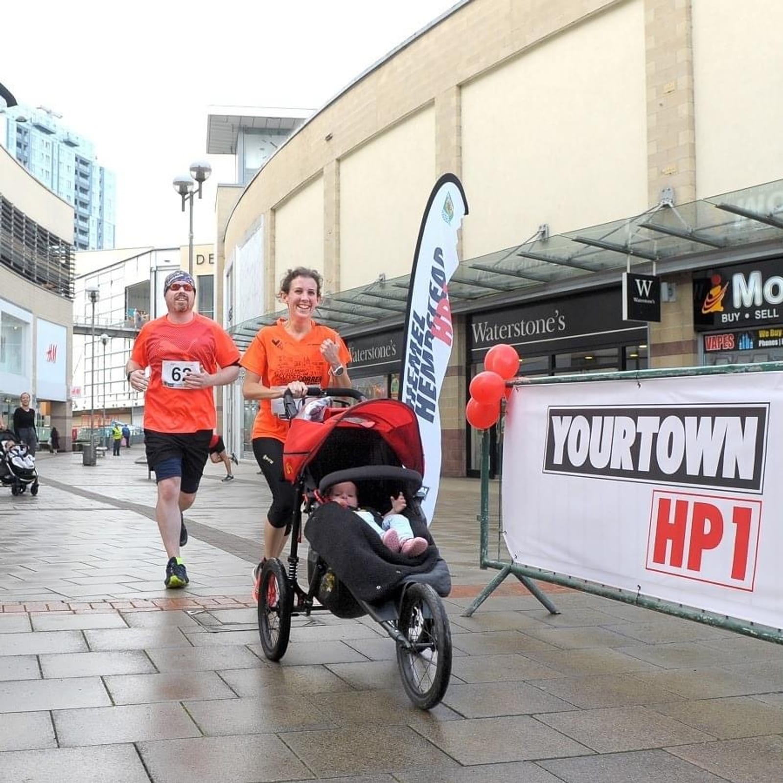 A man and a woman jog in an outdoor race near a shopping area, both wearing orange shirts. The woman pushes a stroller with a baby inside. A banner reading "YOURTOWN HP1" is displayed nearby, and the storefronts of Waterstone's and Mothercare are visible.