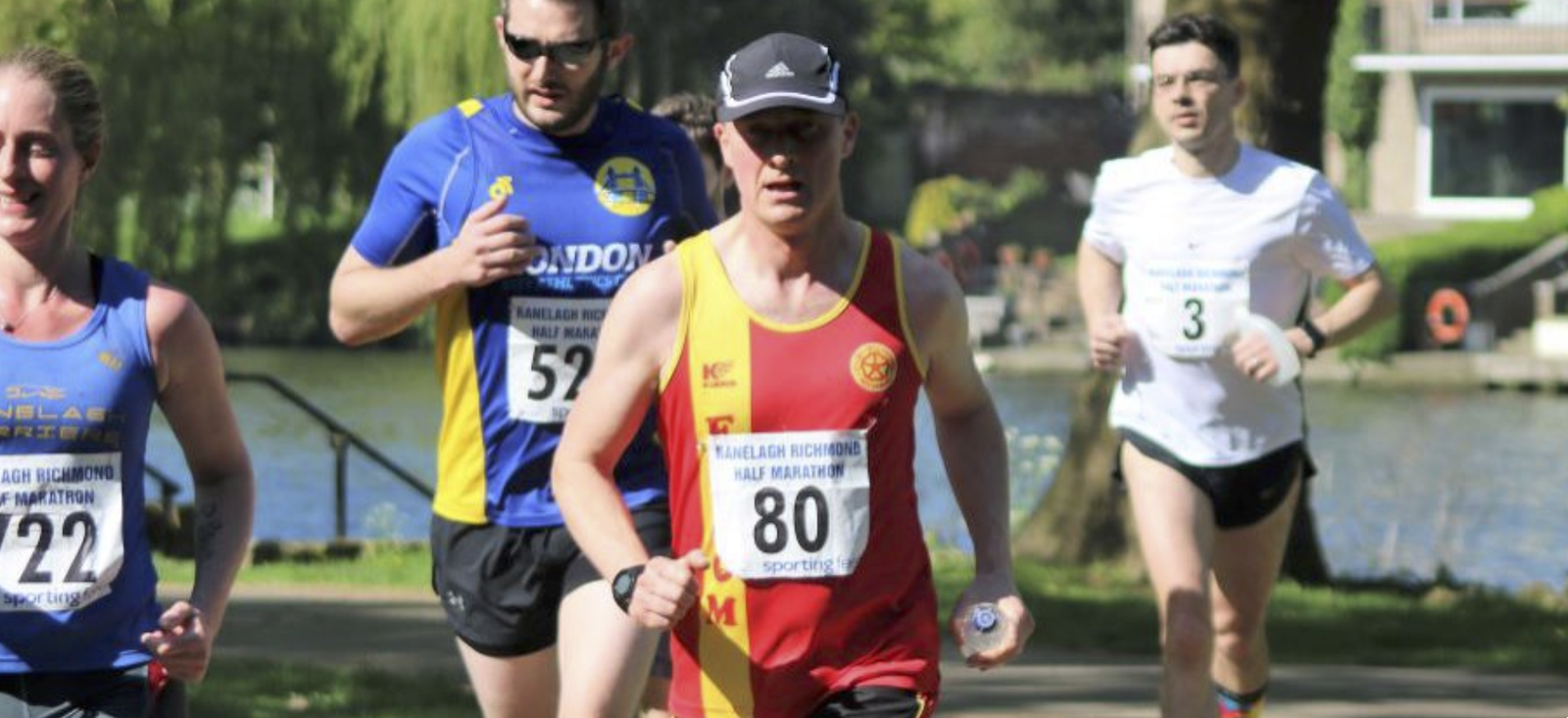Four runners participate in an outdoor race near a body of water and greenery. The runner in the foreground wears a red and yellow outfit with the number 80. Other runners, wearing various outfits and numbers, are visible behind, with one in a blue shirt numbered 52.