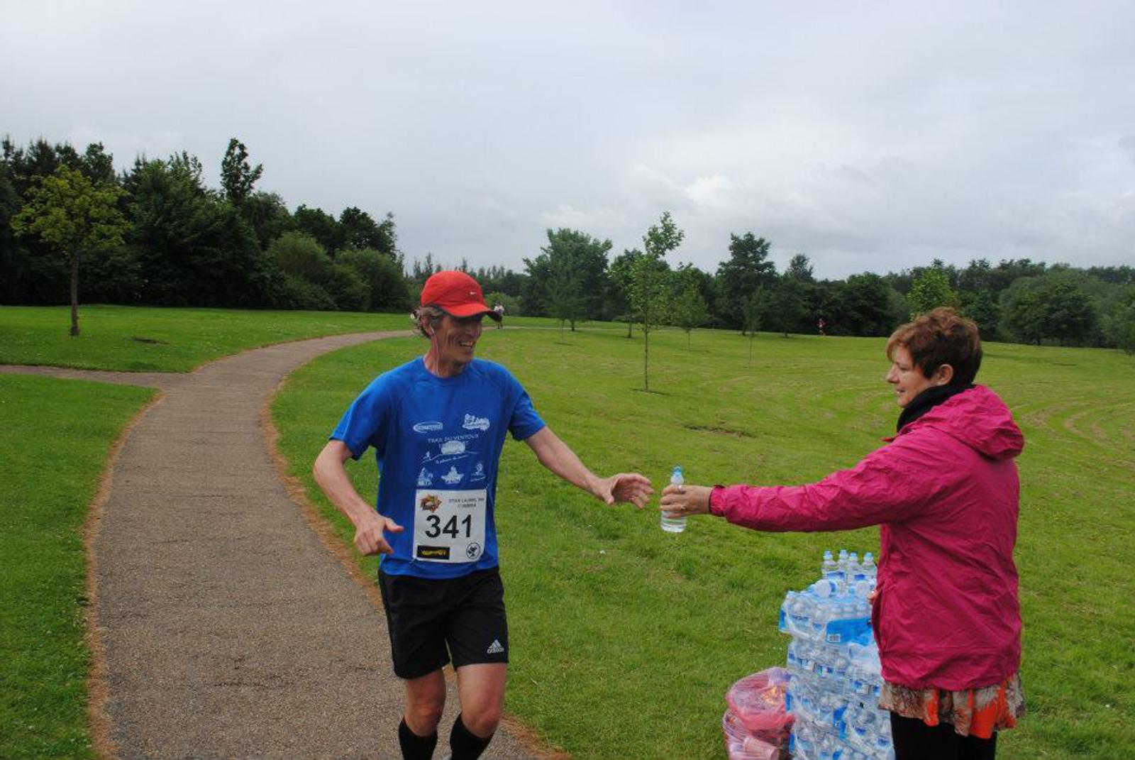 A runner in a blue shirt and red cap approaches a woman in a pink jacket, who is handing him a water bottle. The runner's number is 341. They are on a park path surrounded by green grass and trees.