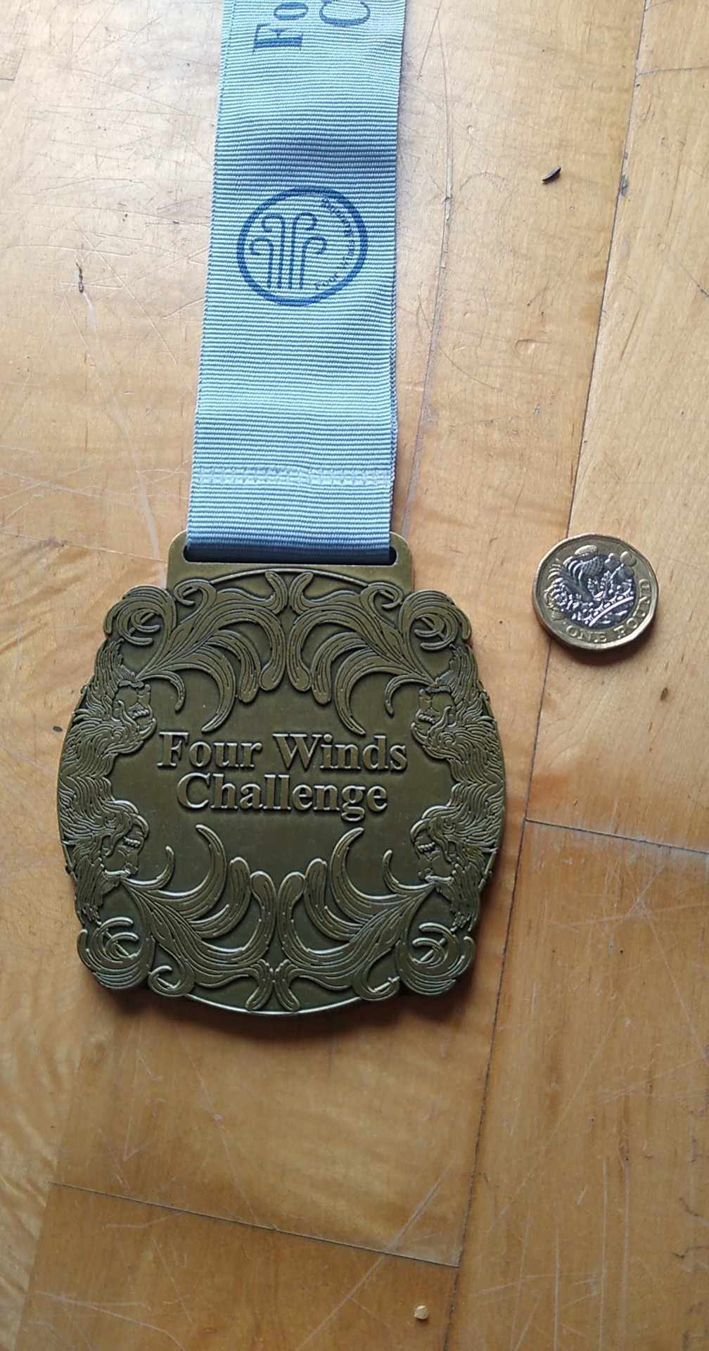 A large gold medal with an intricately designed border and the text "Four Winds Challenge" engraved in the center. The medal is attached to a blue ribbon with additional text visible. A small coin is placed next to the medal for size comparison.