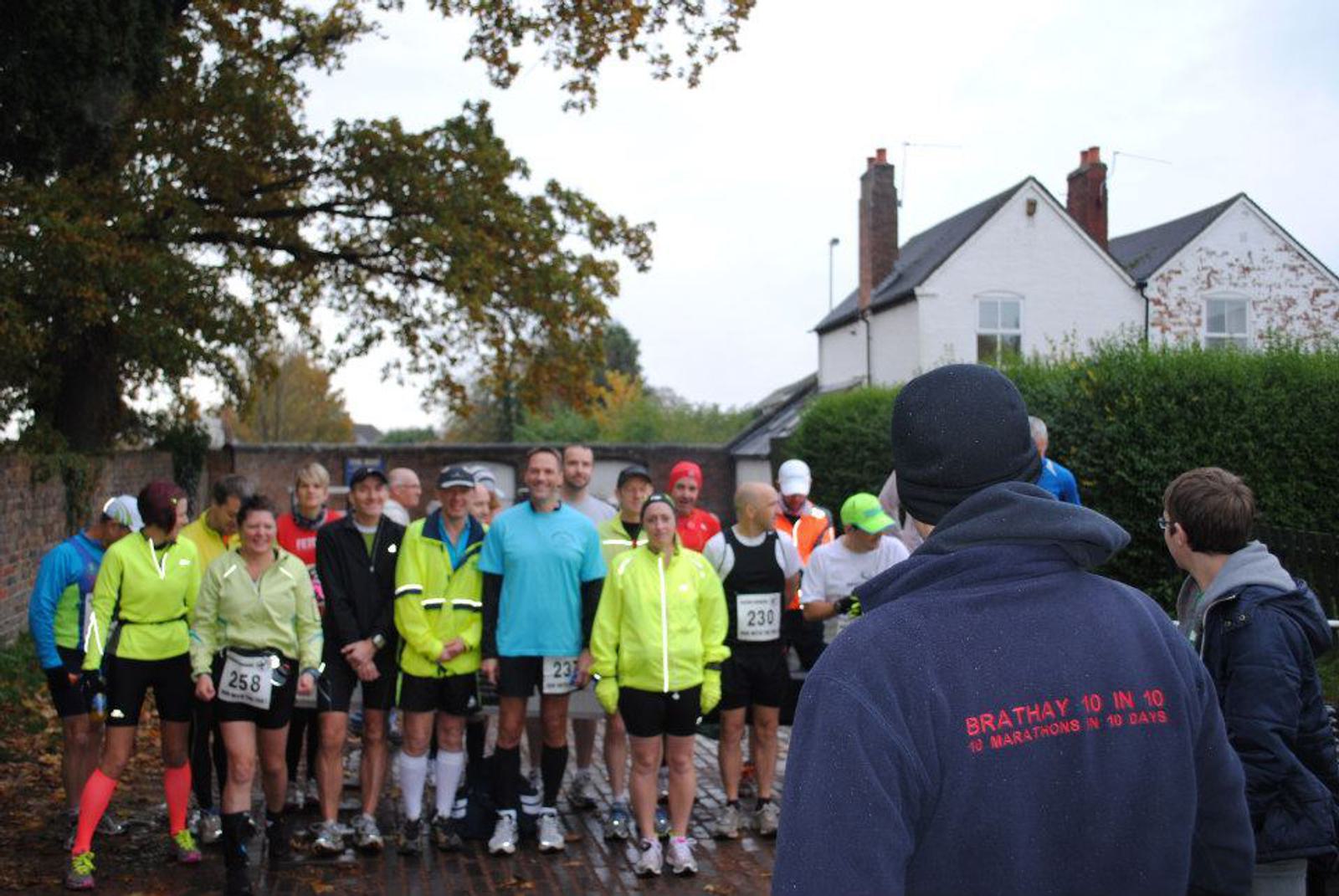 A group of runners wearing athletic gear, standing together on a brick path. Some wear race numbers. Two people, facing them, are dressed in casual clothing and jackets. Houses and trees are visible in the background, under an overcast sky.
