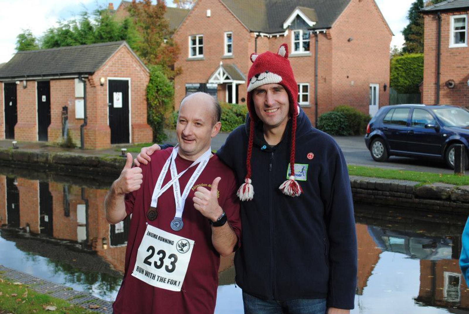 Two people posing outdoors near a canal and brick houses. One is wearing a race bib and medals, giving a thumbs-up. The other is in a quirky fox hat, smiling. A blue car is parked nearby.