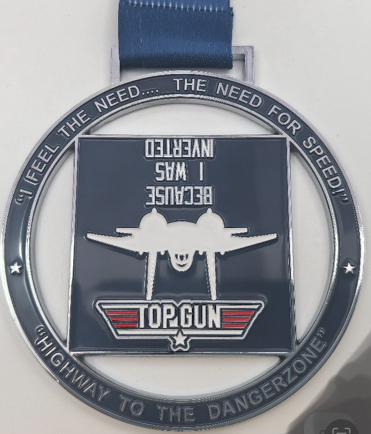 A silver "Top Gun" medal with a blue ribbon. The center features an upside-down fighter jet and the text "BECAUSE I WAS INVERTED." The ring around the medal says, "I FEEL THE NEED... THE NEED FOR SPEED!" and "HIGHWAY TO THE DANGERZONE!.