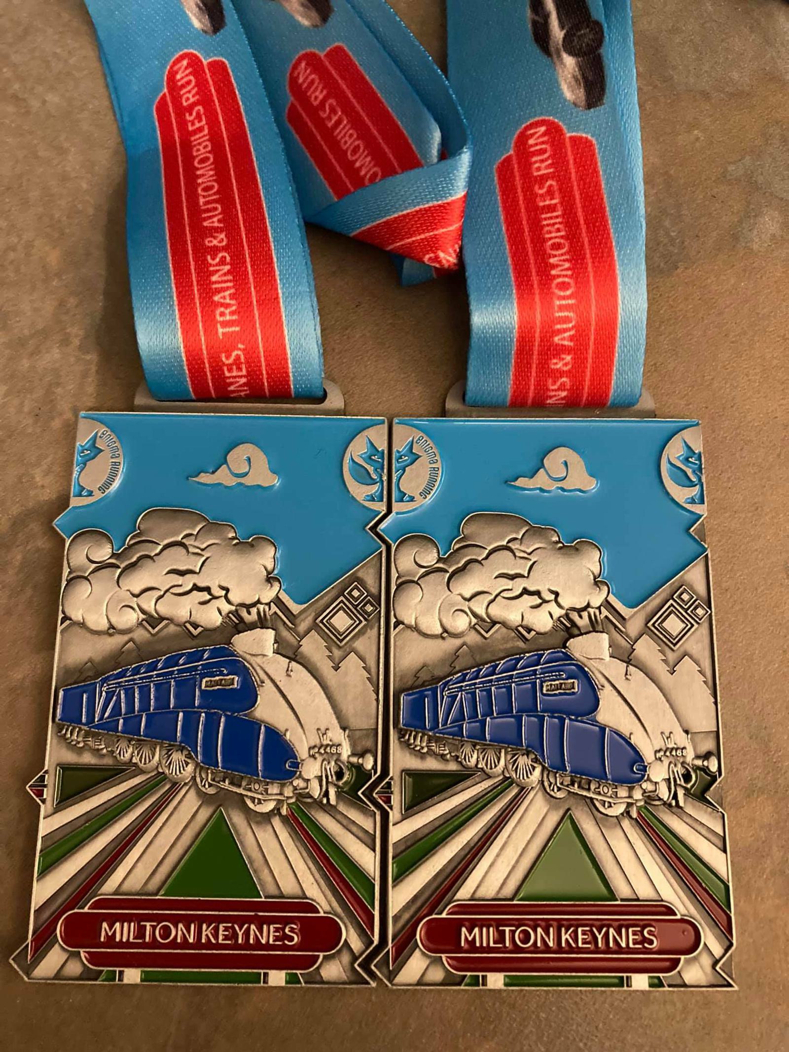 Two medals from the ‘Milton Keynes - Planes, Trains, and Automobiles Run’ are showcased. Each medal features a stylized blue train on tracks, with clouds in the background. The lanyards are blue with red and white text that reads "Planes, Trains & Automobiles Run.
