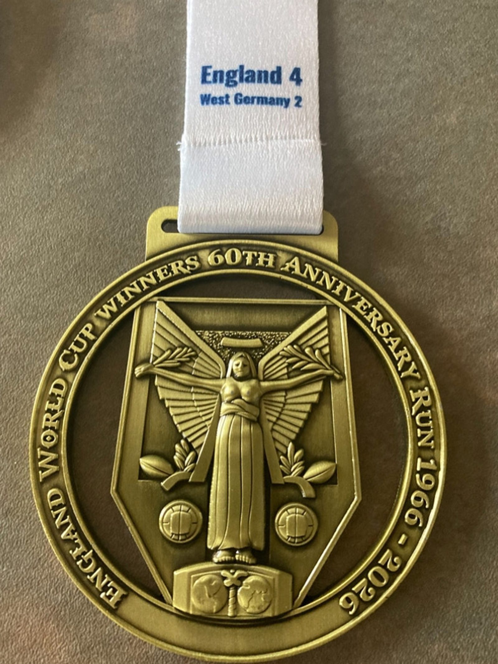 A gold medal with an angel and two footballs, surrounded by text: "England World Cup Winners 60th Anniversary Run 1966-2026." The attached ribbon reads, "England 4 West Germany 2.