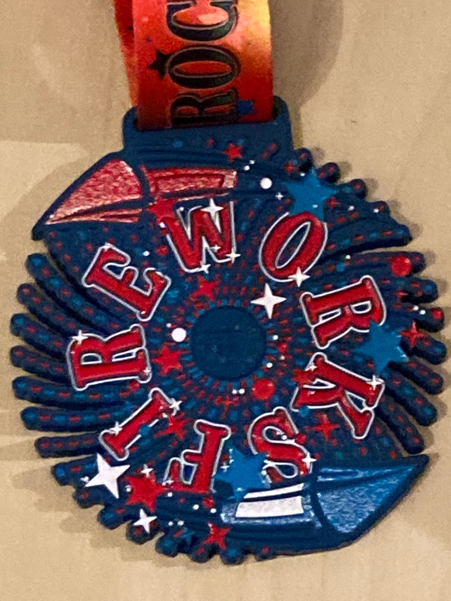 A round medal featuring the word "FIREWORKS" in red and white lettering arranged in a spiral on a blue background with star patterns. Two cartoon-style fireworks are depicted in red and blue. An orange ribbon is attached to the medal.