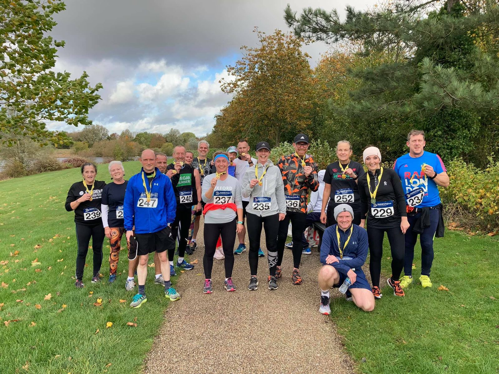 A group of runners stands on a park pathway, posing for a group photo after a race. They wear race bibs, medals, and athletic clothing, with trees and greenery in the background. The sky is partly cloudy, and a few fallen leaves are scattered on the grass.