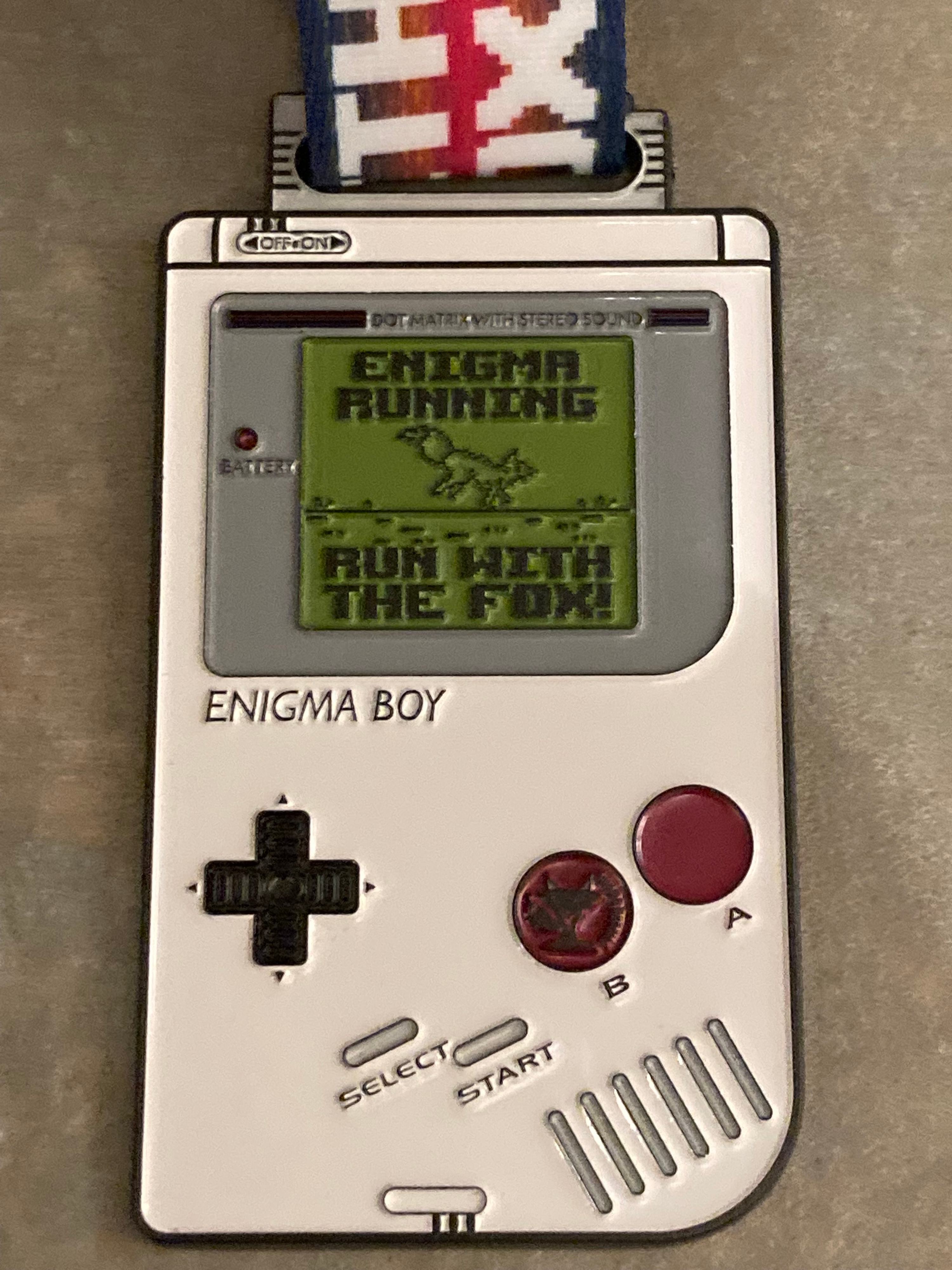 A close-up of a vintage Game Boy-like handheld device resembling a necklace charm. The screen displays the game "ENIGMA RUNNING" with the subtitle "RUN WITH THE FOX!" It has a directional pad, A and B buttons, and a "Select" and "Start" button. "ENIGMA BOY" is written at the bottom.