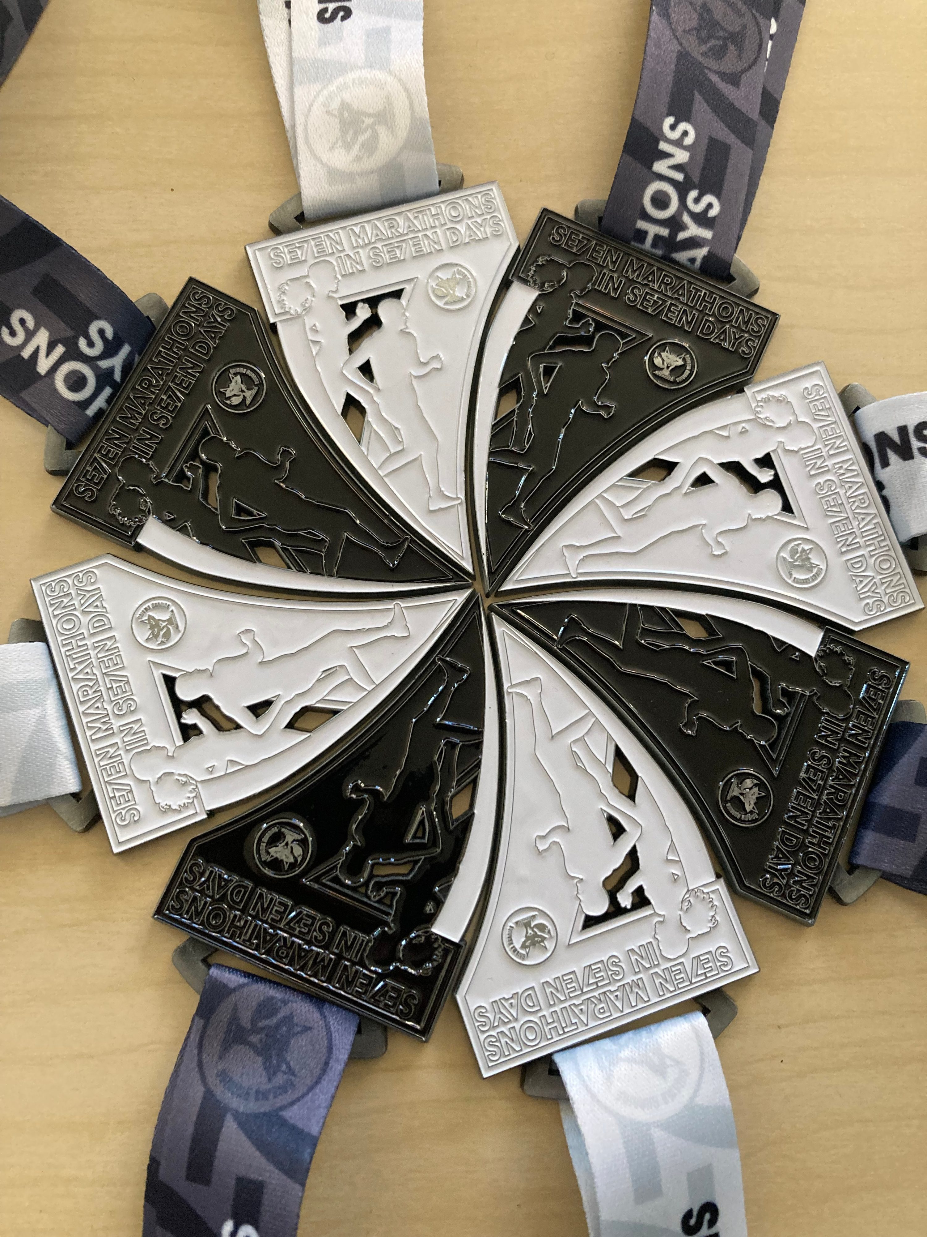 Six black and white marathon medals with running figures create a circular pattern on a light surface. The medals read “Six Marathons in Seven Days” and have matching patterned ribbons.